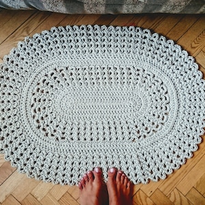 May include: A light gray oval crochet rug with a delicate lace pattern. The rug is laid out on a wooden floor.