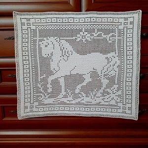 May include: A square, off-white crocheted doily featuring a detailed horse design in the center. The doily has a decorative border with a repeating pattern of small holes. The handmade textile is placed on a dark wooden surface.