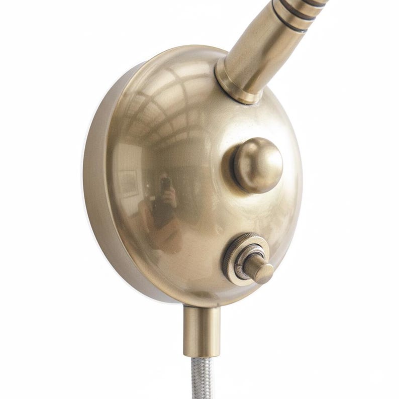 May include: Close-up of a brass-colored wall-mounted lamp with a flexible gooseneck. The lamp features a round head with a button and a switch. The flexible neck is connected to a silver braided cable.