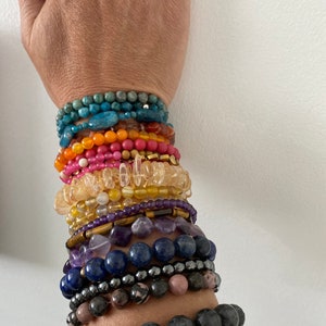 May include: A stack of colorful gemstone bracelets on a person's wrist. The bracelets are made of various stones, including turquoise, carnelian, citrine, amethyst, lapis lazuli, hematite, and rose quartz.