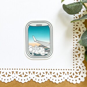 May include: A sticker of an airplane wing seen through an airplane window. The window is outlined in white and the sticker is on a white background.