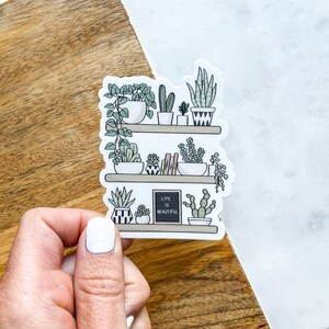 May include: A sticker featuring a hand-drawn illustration of a shelf with potted plants and a small chalkboard sign that reads "Life is Beautiful".