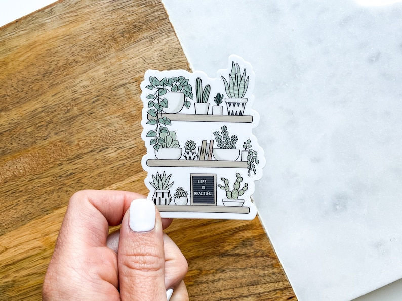 May include: A sticker featuring a hand-drawn illustration of a shelf with potted plants and a small chalkboard sign that reads "Life is Beautiful".