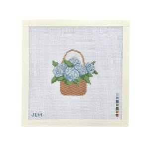 May include: A square needlepoint canvas featuring a cross-stitched design of a brown woven basket filled with light blue hydrangea flowers and green leaves. The canvas includes a color palette guide on the right side and the letters JLM in blue.