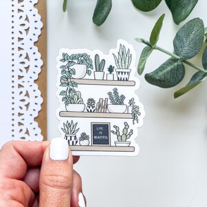 May include: A sticker featuring a hand-drawn illustration of a shelf with potted plants. The shelf has three tiers and is filled with various plants, including succulents, cacti, and ferns. The sticker also includes a small chalkboard sign that reads "Life is Beautiful".