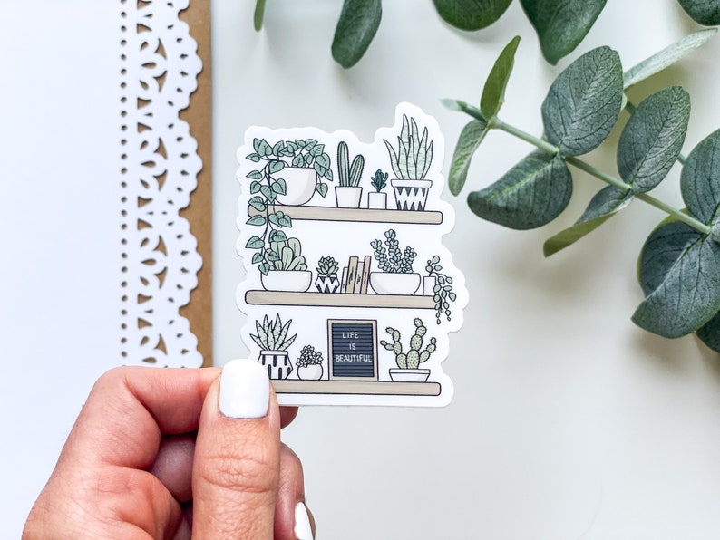 May include: A sticker featuring a hand-drawn illustration of a shelf with potted plants. The shelf has three tiers and is filled with various plants, including succulents, cacti, and ferns. The sticker also includes a small chalkboard sign that reads "Life is Beautiful".