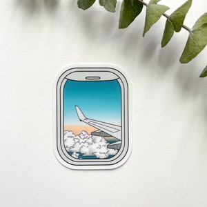May include: A sticker of an airplane window with a view of clouds and a wing of the plane against a blue sky.