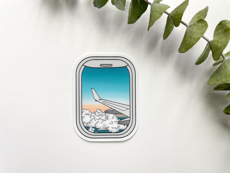 May include: A sticker of an airplane window with a view of clouds and a wing of the plane against a blue sky.