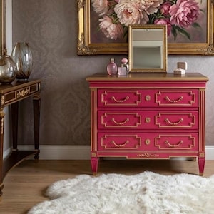 May include: A vibrant pink dresser with gold trim and hardware. The three-drawer chest features a rectangular design with ornate detailing. A gold-framed mirror and perfume bottles sit atop the dresser, with a large floral painting in the background.