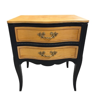 May include: A two-drawer nightstand with a light wood top and drawer fronts. The frame is black with ornate brass drawer pulls. The nightstand has curved legs and a decorative trim between the drawers. The overall style is French Provincial.