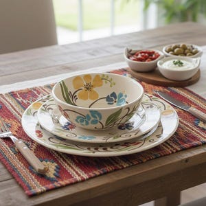 May include: A floral-patterned ceramic dinnerware set, including a bowl and two plates, sits on a patterned placemat. A fork and knife with wooden handles are on the table. Small bowls of food are in the background.