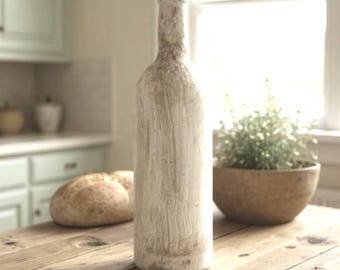 May include: A tall, off-white decorative bottle with a textured surface and a cork stopper. The bottle is displayed on a rustic wooden table, alongside a wooden bowl with greenery and a loaf of bread. The scene is set in a kitchen.