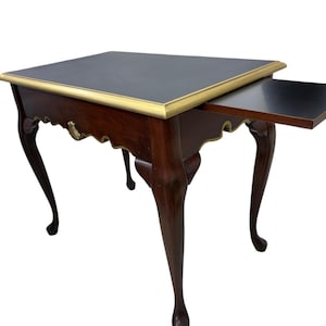 May include: A dark wood side table with a black top and a pull-out section. The table has a gold trim around the top and decorative gold accents. The table's legs are curved. The table measures approximately 24 inches wide, 20 inches deep, and 28 inches high.