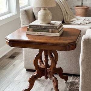 May include: A wooden side table with a square top and a central pedestal base featuring four curved legs on casters. A beige lamp sits on a stack of three books titled Design, History on My Hertor, and Natural History on the table surface.