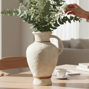 May include: A cream-colored, textured vase with a handle and a rope detail around the neck and base. The vase holds a bouquet of white flowers and green foliage. The vase is on a light-colored wooden table.