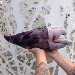 May include: A plush toy shaped like a sea creature, held up by two hands. The toy has a dark purple and gray body with a lighter gray head and a red mouth. The background is a white wall with mirrored cutouts.