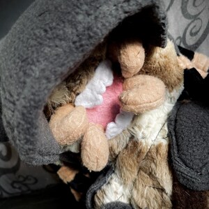 Op de afbeelding: Een pluche speelgoed met een bruin en wit gestreepte vacht, met een zwarte capuchonjas. Het speelgoed heeft een grote, open mond met witte tanden en een roze tong.