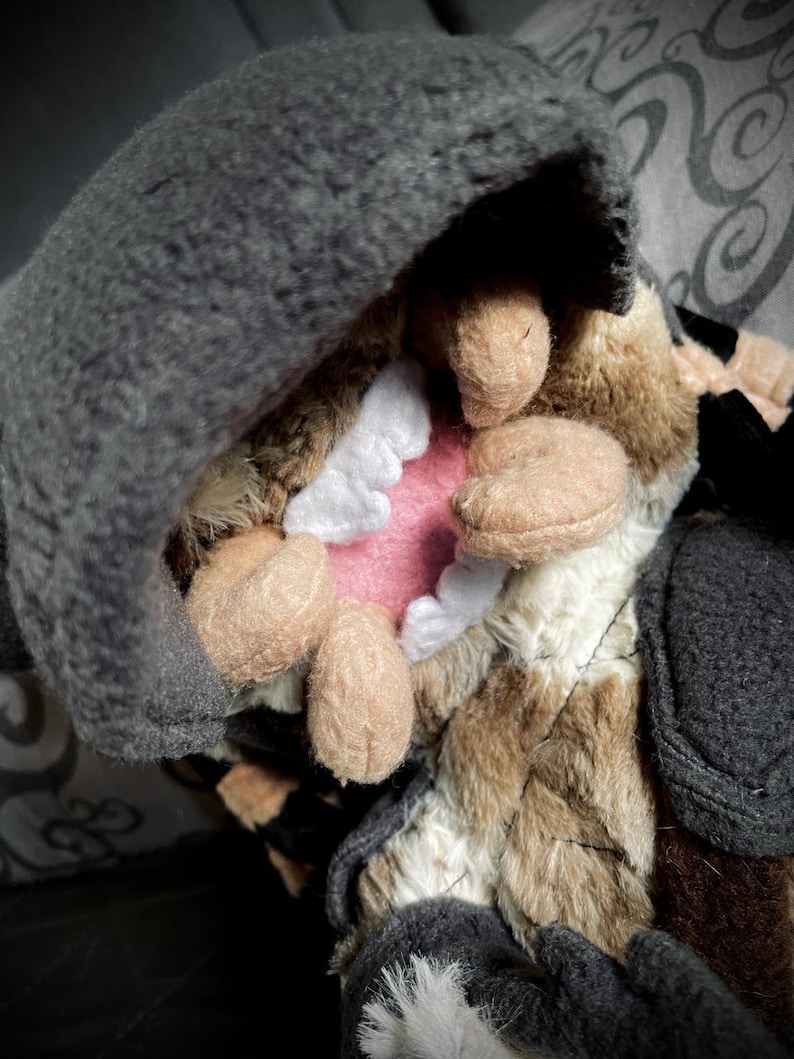 Op de afbeelding: Een pluche speelgoed met een bruin en wit gestreepte vacht, met een zwarte capuchonjas. Het speelgoed heeft een grote, open mond met witte tanden en een roze tong.