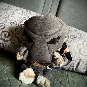 May include: A plush toy of a Predator character, wearing a black and grey mask, sitting on a green bedspread.