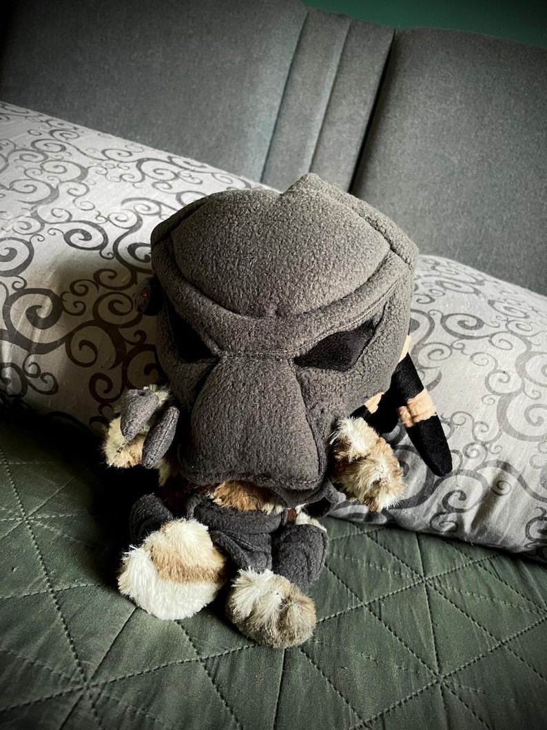 Op de afbeelding: Een pluche speelgoed van een Predator-personage, met een zwart en grijs masker, zittend op een groene bedsprei.