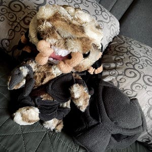 Op de afbeelding: Een pluche speelgoed met een bruin en zwart camouflagepatroon, een grote, tandenrijke grijns en zwarte klauwen. Het speelgoed ligt op een grijs en wit gepatroneerd kussen.