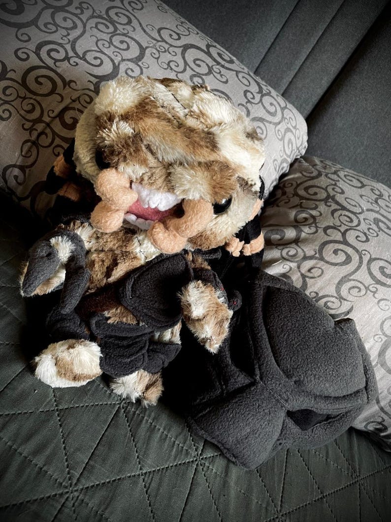 Op de afbeelding: Een pluche speelgoed met een bruin en zwart camouflagepatroon, een grote, tandenrijke grijns en zwarte klauwen. Het speelgoed ligt op een grijs en wit gepatroneerd kussen.