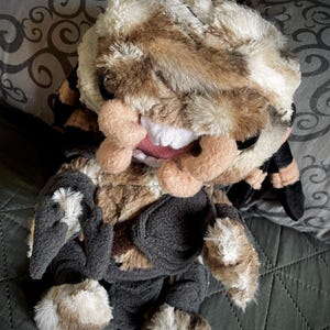 Op de afbeelding: Een pluche speelgoed met bruin en wit bont, dat een zwarte outfit draagt. Het speelgoed heeft een brede, tanden grijns en een ondeugende uitdrukking.