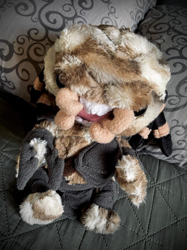 Op de afbeelding: Een pluche speelgoed met bruin en wit bont, dat een zwarte outfit draagt. Het speelgoed heeft een brede, tanden grijns en een ondeugende uitdrukking.