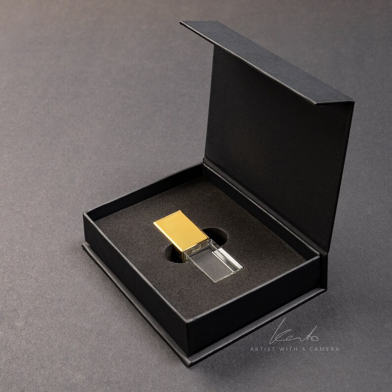 May include: A clear crystal USB drive with a gold top is displayed in a black gift box. The box is open and the drive is sitting in the center of the box.
