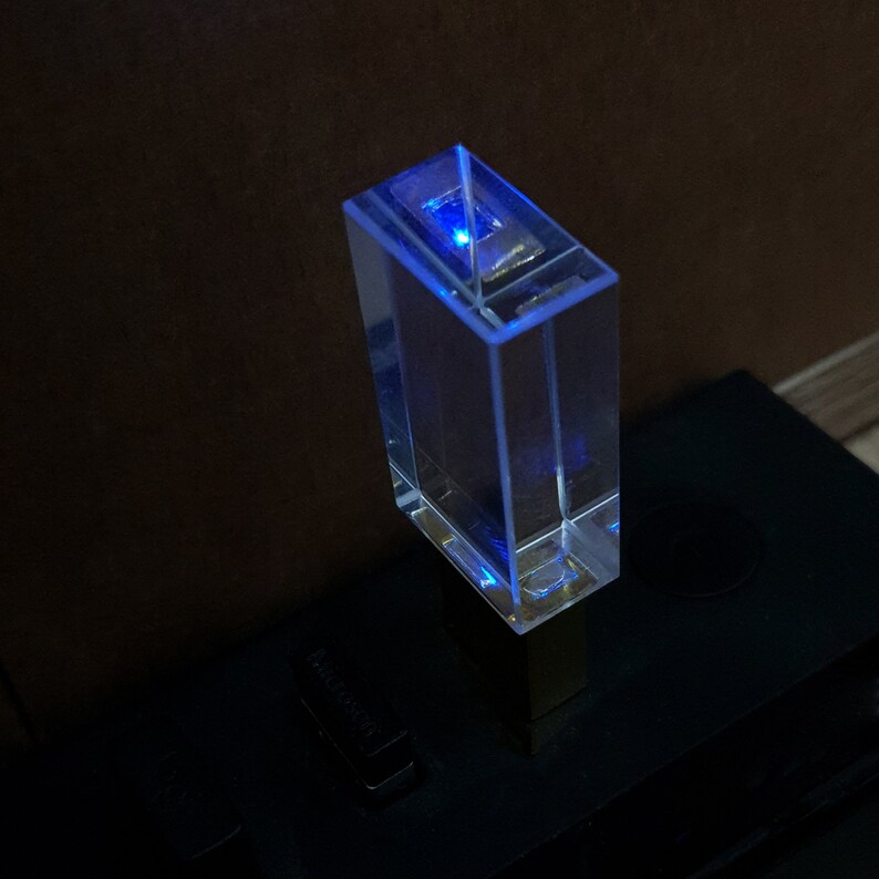 May include: A clear glass USB drive with a blue light shining from the top. The drive is plugged into a black USB port.