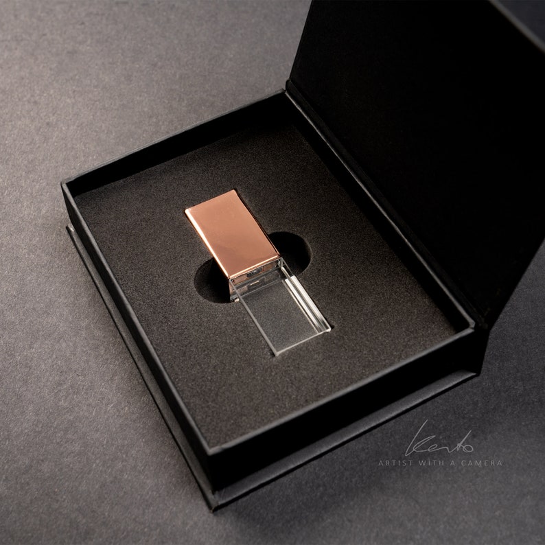 May include: A clear crystal USB drive with a rose gold top in a black velvet box. The box is open and the drive is sitting inside.