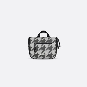 May include: A rectangular toiletry bag featuring a black and white houndstooth pattern. The bag includes a top handle, a horizontal zippered front pocket, and a side zipper closure. The brand name SHERPANI is printed in small yellow letters on the front.