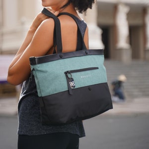 A curly haired model is facing away from the camera, looking over her left shoulder and smiling. She is wearing a gray tank top and black leggings. She is holding Sherpani's Anti-Theft Tote, the Cali AT in Teal, over her shoulder.