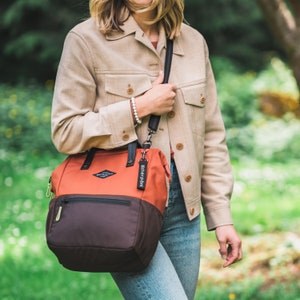 A blonde woman stands outside in a park. She is wearing a tan jacket and jeans. She carries Sherpani three-in-one bag, the Dispatch in Clay, as a crossbody.