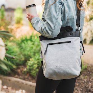 Close up view of a blonde haired model standing outside holding a coffee. She is wearing a jean jacket, black leggings and Sherpani Anti-Theft bag, the Geo AT in Sterling, as a crossbody.
