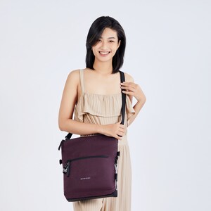 Close up view of a dark haired model facing the camera and smiling. She is wearing a tan pinstriped jumpsuit. She holds Sherpani Anti-Theft bag, the Geo AT in Merlot as a crossbody.