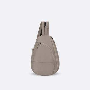May include: A teardrop-shaped sling backpack in a solid light beige color. The bag features a smooth exterior, a single adjustable shoulder strap, a curved front zippered pocket, and a main vertical zippered compartment with matching beige zipper pulls.