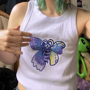Op de afbeelding: Een wit geribbeld hemdje met een centrale afbeelding van een gestileerde mot of vlinder. De illustratie van het insect is uitgevoerd in aquarel-stijl met gemengde tinten paars, blauw en geel, omlijnd met een dikke, donkerpaarse inktlijn.