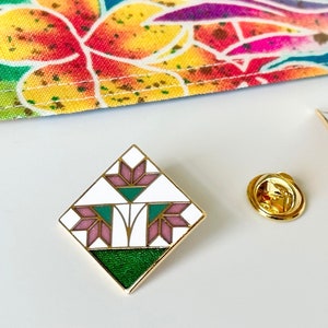 May include: A gold-toned enamel pin with a geometric floral design in white, green, and purple. A matching pin with a pin back and a gold-colored pin back are also present. A colorful floral fabric is in the background.