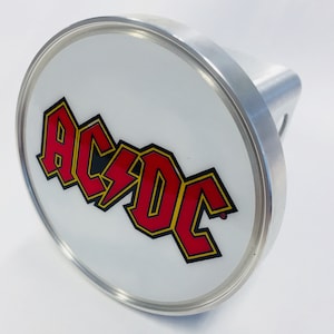 May include: A silver hitch cover with a white background and a red and yellow AC/DC logo.