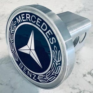 May include: A silver hitch cover with a black and white Mercedes-Benz logo. The logo features a three-pointed star inside a circle with the words "Mercedes-Benz" around the outside.