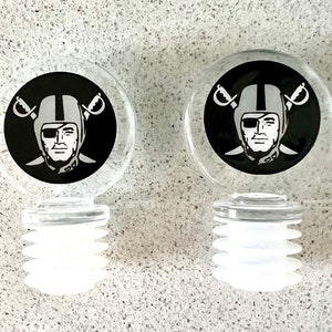 May include: Two clear glass bottle stoppers with a black and white Raiders logo. The logo features a player wearing a helmet with a black eye patch and a white skull and crossbones.