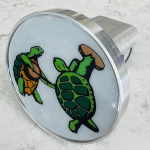 May include: A silver-colored metal hitch cover with a white background featuring two cartoon turtles. One turtle plays a banjo, and the other holds a tambourine. The turtles are green and orange.