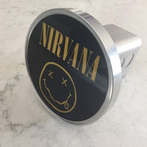 May include: A black and gold metal hitch cover with the word "NIRVANA" and a smiley face with an X for eyes and a tongue sticking out.