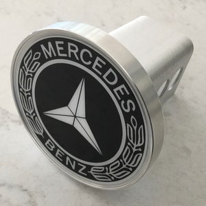 May include: Silver hitch cover with a black and white Mercedes-Benz logo. The logo features a three-pointed star and the words "Mercedes-Benz" around the star.