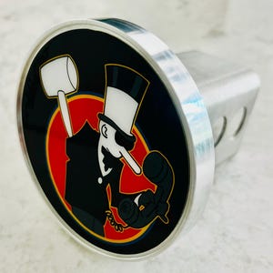 May include: A black and white cartoon character with a top hat and a long nose holding a phone. The character is wearing a black suit and a white shirt. The character is standing in front of a red circle with a yellow outline. The image is on a silver hitch cover.