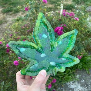 May include: A green resin leaf-shaped tray with a textured surface and embedded white stones. The tray has a glossy finish and is held in front of a background of pink flowers and green foliage.