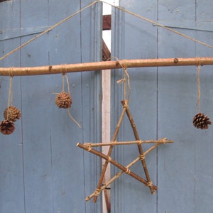May include: A rustic wooden star ornament hanging from a branch with pine cones on twine. The star is made of twigs and the pine cones are natural brown.