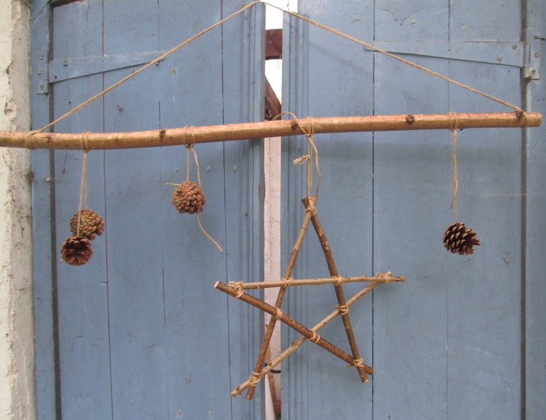 May include: A rustic wooden star ornament hanging from a branch with pine cones on twine. The star is made of twigs and the pine cones are natural brown.