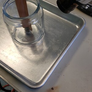 May include: A heat gun is being used to heat a glass jar with a brown paper strip inside. The jar is sitting on a silver baking sheet.